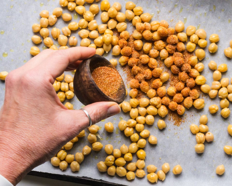 Spices are being poured over chickpeas in a pan lined with parchment paper.