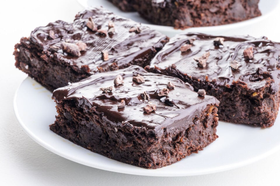 A plate of gooey frosted date brownies are topped with cacoa nibs and sprinkles of flaky sea salt.