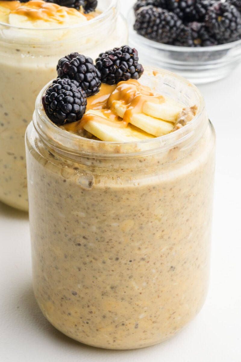 Oatmeal jar topped with blackberries, banana slices, and drizzled peanut butter, with another jar in the background.