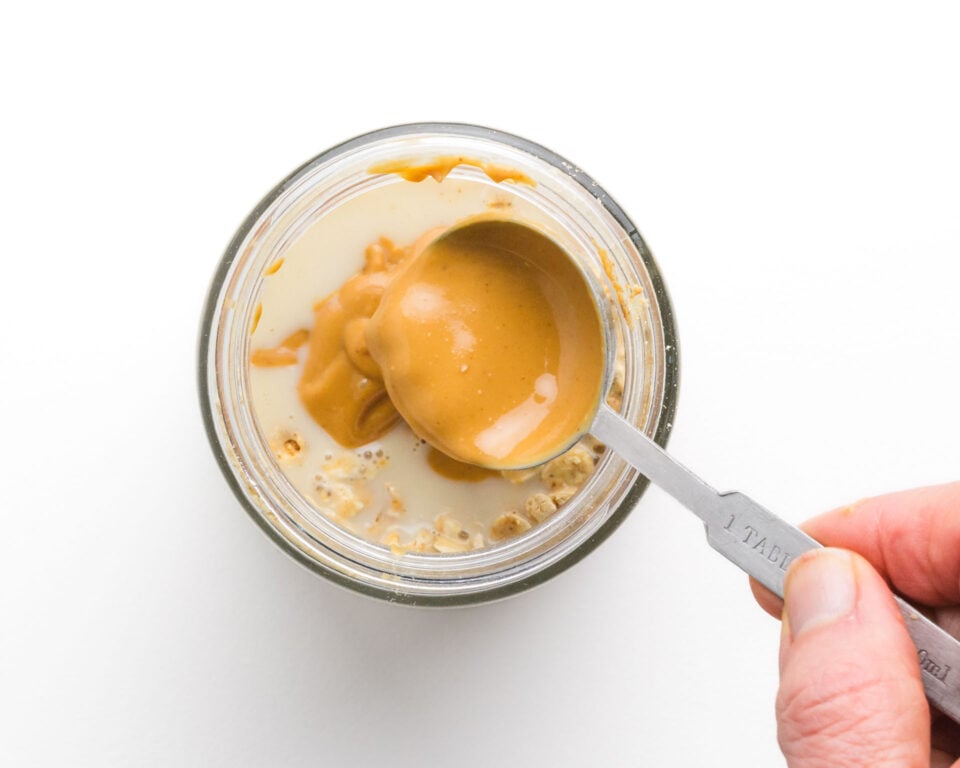 Spoon holding peanut butter above a jar containing milk and granola.