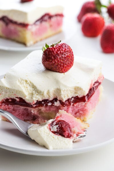 Strawberry shortcake slice topped with fresh strawberry and whipped cream, served with a fork on a white plate.