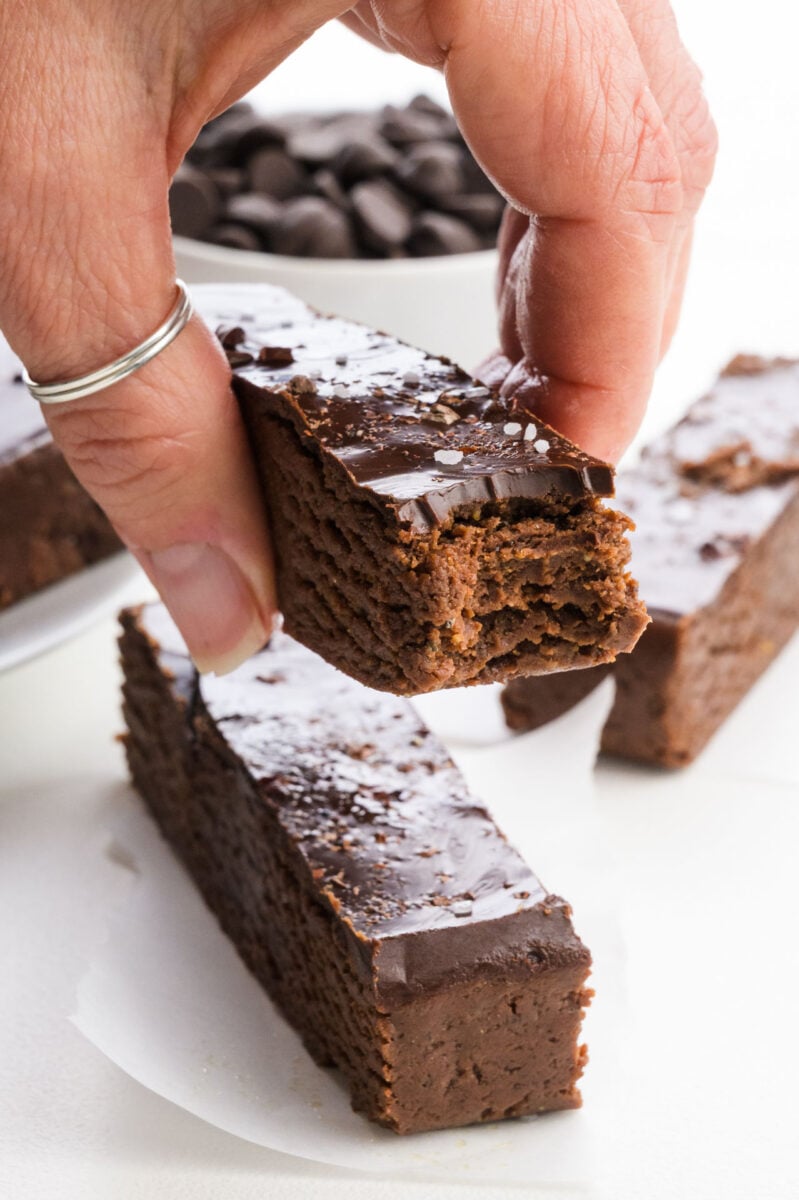 A hand holding a chocolate protein bar with a shiny topping, surrounded by other chocolate bars and chocolate chips.