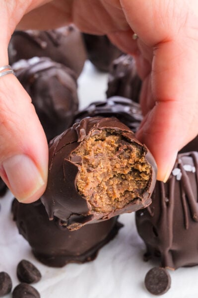 A hand holding a chocolate-covered treat with a bite taken out, revealing a textured filling inside.